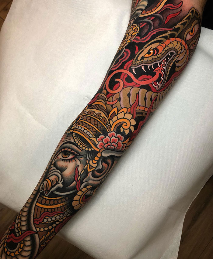  A detailed full-sleeve tattoo featuring a vibrant traditional design with intricate patterns, showcasing a fierce serpent and a deity with elaborate headgear. Tattoo ideas for men wanting bold and mythological art with striking reds, golds, and detailed black outlines.