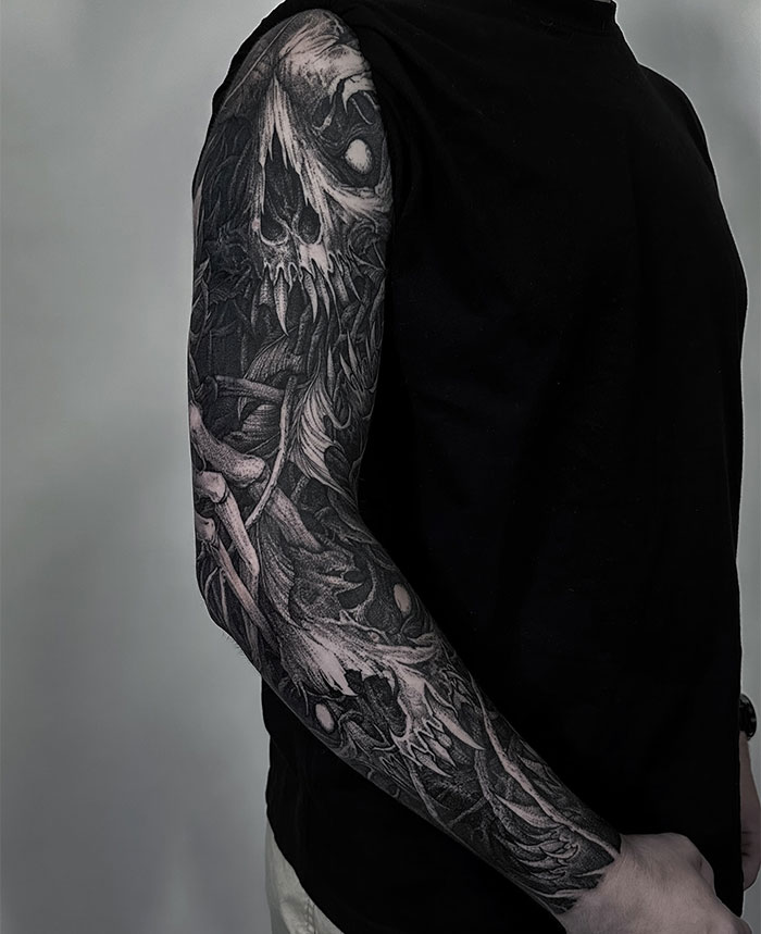 A detailed black and gray full-sleeve tattoo featuring a haunting skull with sharp teeth and intricate shadows, conveying a dark and intense design. Tattoo ideas for men seeking bold, eerie, and highly detailed arm art with a gothic and mysterious theme.