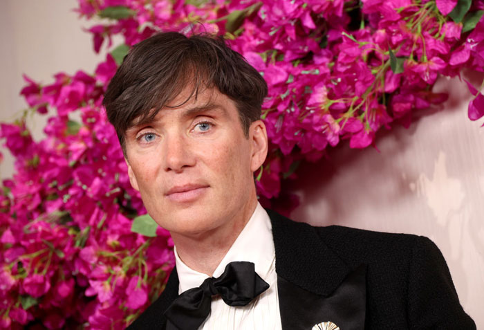  A B list celebrity, Cillian Murphy, is posing in a black tuxedo with a bow tie in front of vibrant pink flowers at an indoor event.