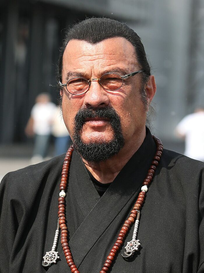 Actor in black attire and glasses, standing outdoors with a focused expression.
