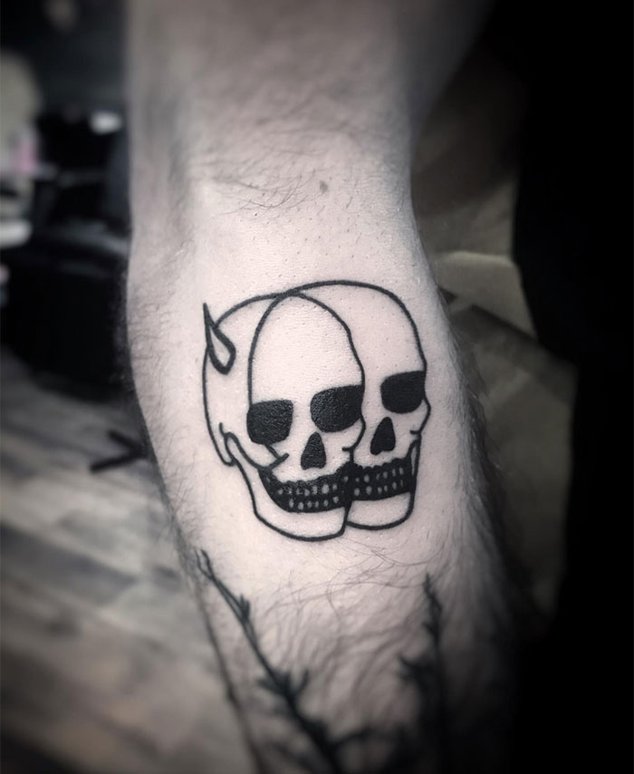 A black ink tattoo of two conjoined skulls, one with a small horn, creating a duality theme of good and evil. A minimalist yet bold tattoo idea for men.
