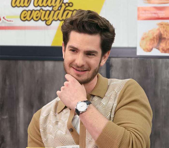 Andrew Garfield smiling, wearing a brown cardigan and a watch, embodiment of a masterclass in seduction.