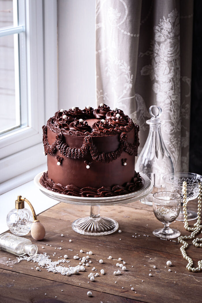 Elegant chocolate cake on a glass stand, celebrating Tiptree Cake Awards, with decorative curtain backdrop.