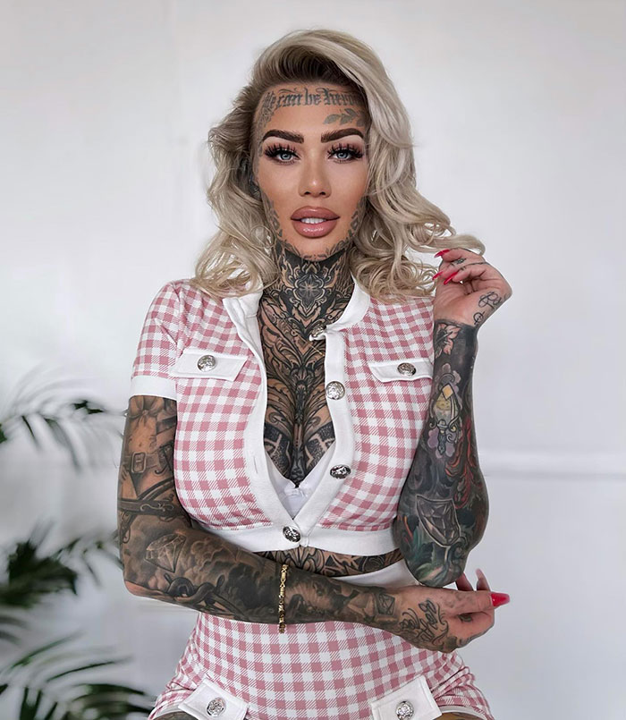 A woman with face tattoos wearing a checkered outfit stands confidently in front of a neutral background.