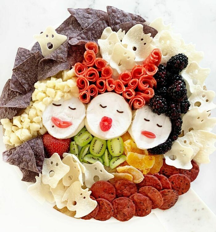 Halloween snacks arranged as three witch faces with various fruits, chips, and ghost-shaped crackers on a platter.