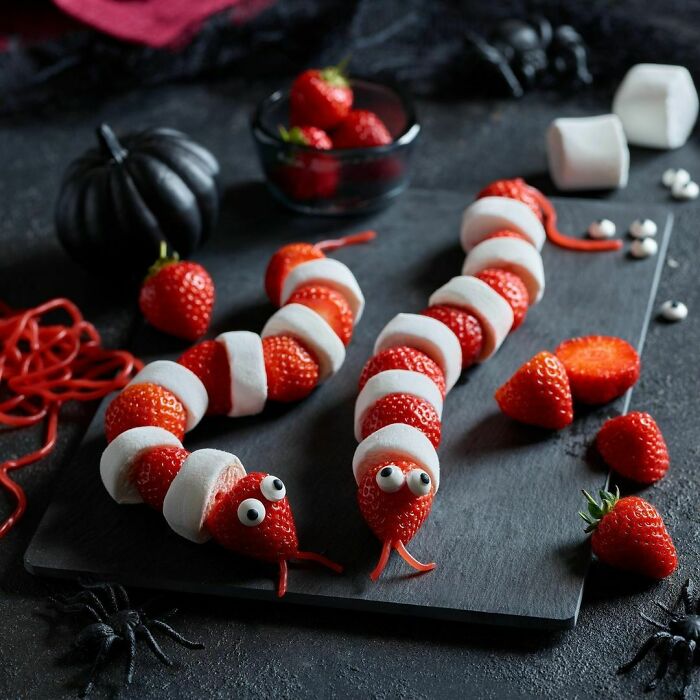 Halloween snacks shaped like snakes with strawberries, marshmallows, and candy eyes on a black serving board.