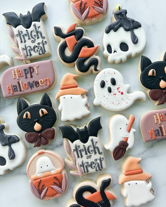 Halloween snacks decorated as ghost, black cat, skull, and bat cookies with intricate icing designs on a white marble background.