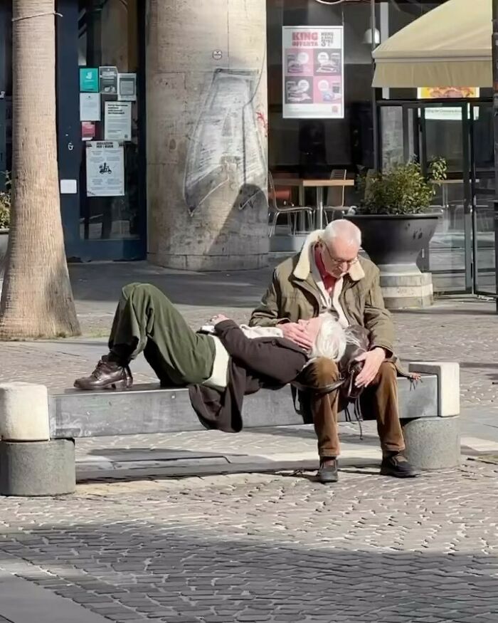 Elderly couple sharing a tender moment on a bench in a sunny urban setting, brightening the day with warmth.