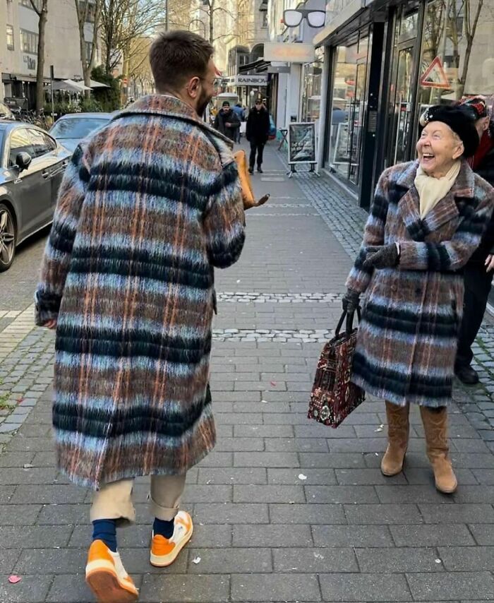 Man and elderly woman wearing matching plaid coats, smiling on a city sidewalk in a delightfully random photo.