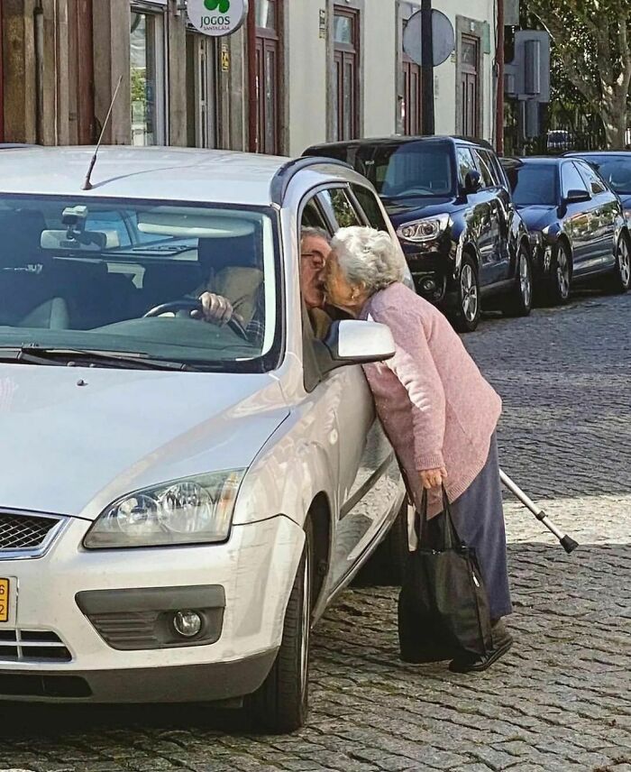 Elderly woman leaning into a car window to kiss an elderly man, a heartwarming delightfully random photo on a street.