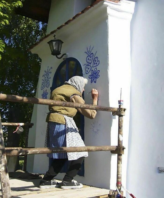 Elderly woman painting blue floral and heart designs on white wall, adding delightfully random art outdoors.