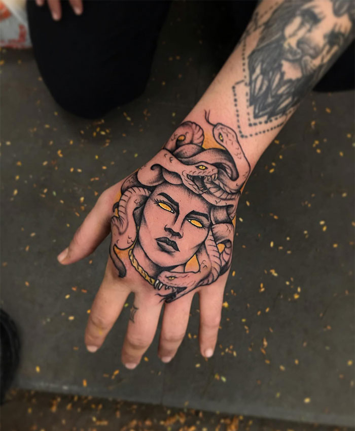 Medusa tattoo on hand with yellow eyes and snakes, shaded in dark and golden tones, symbolizing beauty and power.