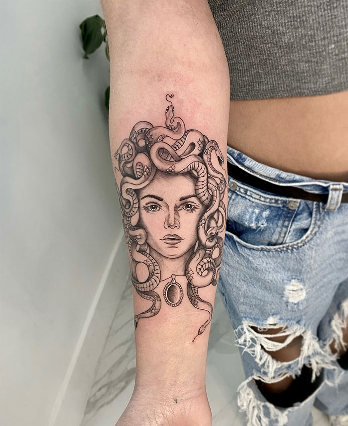 Black and grey Medusa tattoo on forearm, showing a woman's face entwined with snakes and a pendant, symbolizing beauty and defiance.