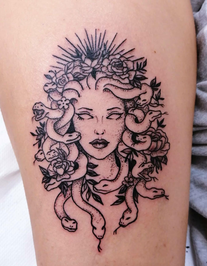 Black and grey Medusa tattoo with blank-eyed woman's face, surrounded by snakes and flowers, rays above her head.
