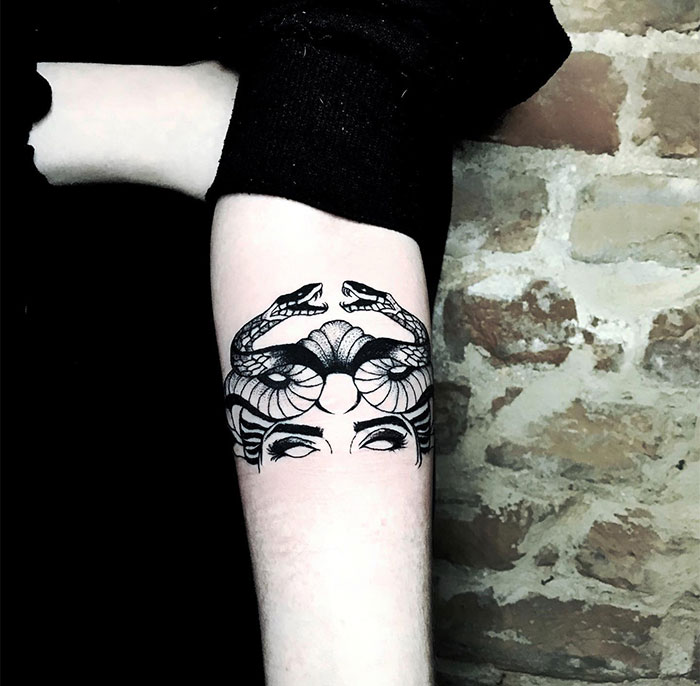 Minimalistic Medusa tattoo on forearm, showing upper face with intertwined snakes, symbolizing beauty and defiance.