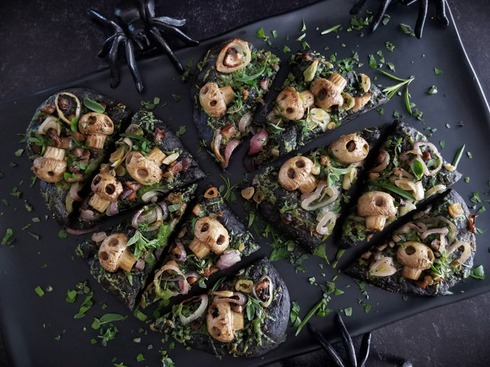 Halloween snacks with black pizza crust topped with mushrooms and herbs on a black platter with decorative spider hands.