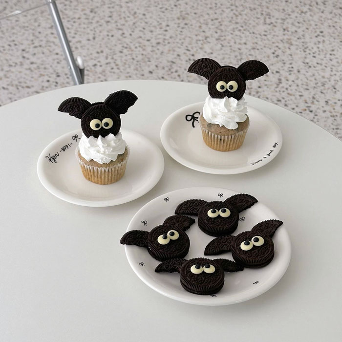 Halloween snacks featuring cupcakes and cookies decorated as bats with Oreo cookies and candy eyes on plates.