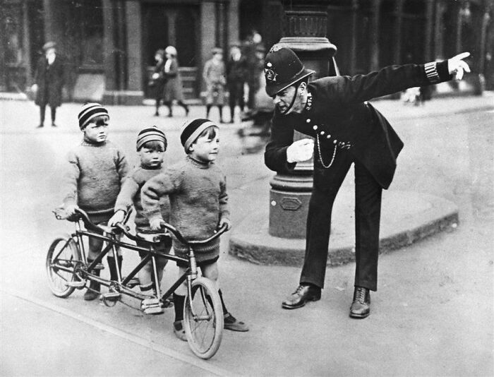 London In The 1920s. A Policeman In A London Street Giving Directions To The Three Children On A Bicycle. The Bicycle Is Specially Made For Three Persons