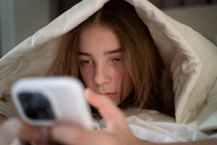 A girl under a blanket looks at a smartphone screen, appearing contemplative.