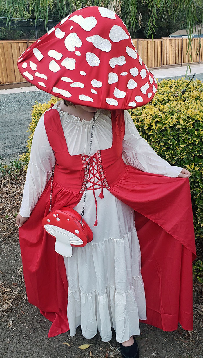 Child wearing a creative Halloween costume with a red and white mushroom theme outdoors near greenery and a wooden fence.