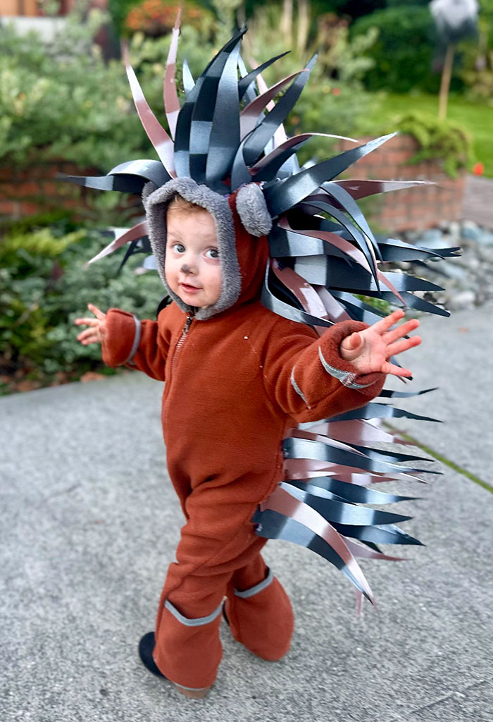 Toddler in a creative kids' Halloween costume with spiky headpiece and brown jumpsuit outdoors on a sidewalk.