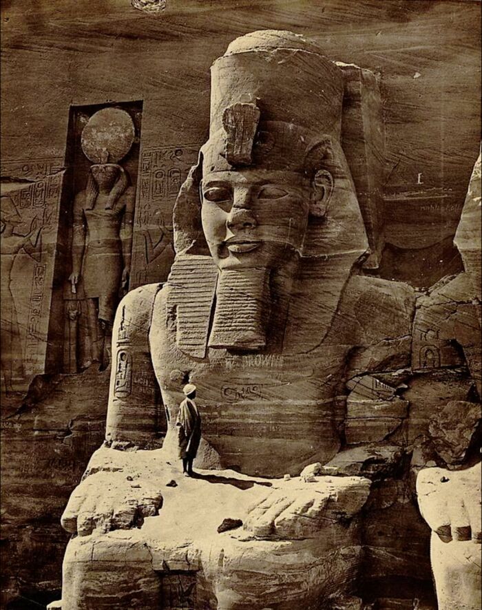 A Photo Of A Man Standing On The Lap Of A Colossal Statue Of Ramses II In 1856, Emphasizing The Monument’s Immense Scale