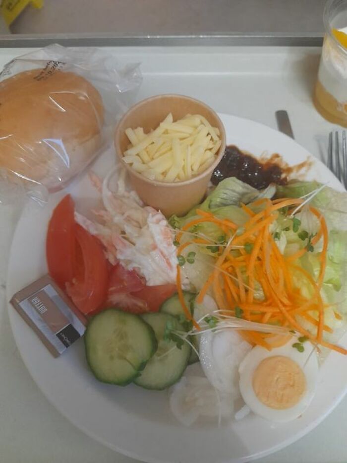 Hospital food example featuring a salad with cucumber, tomato, boiled egg, shredded cheese, and a bread roll on a tray.