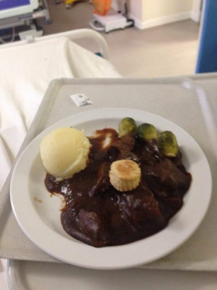Hospital food example featuring mashed potatoes, meat with brown sauce, Brussels sprouts, and a small corn piece on a white plate.