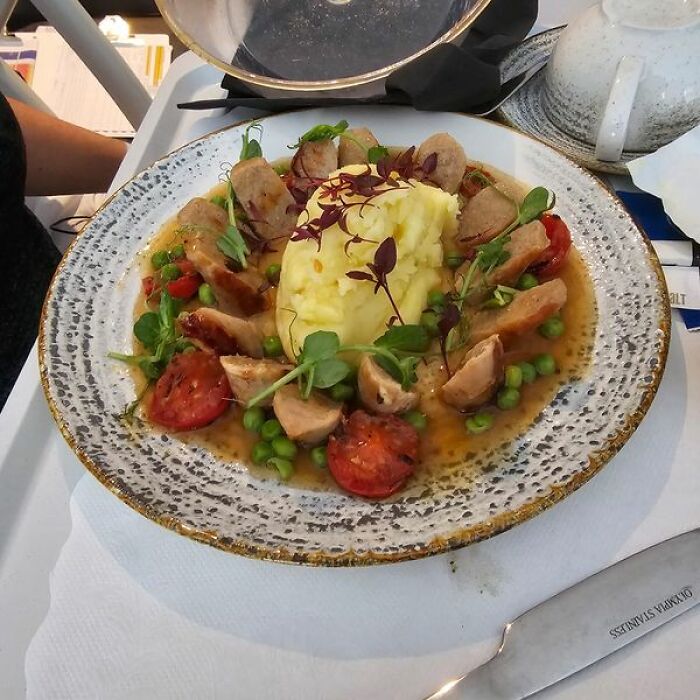 Hospital food example featuring mashed potatoes, sausage slices, peas, tomatoes, and garnishes served on a decorative plate.