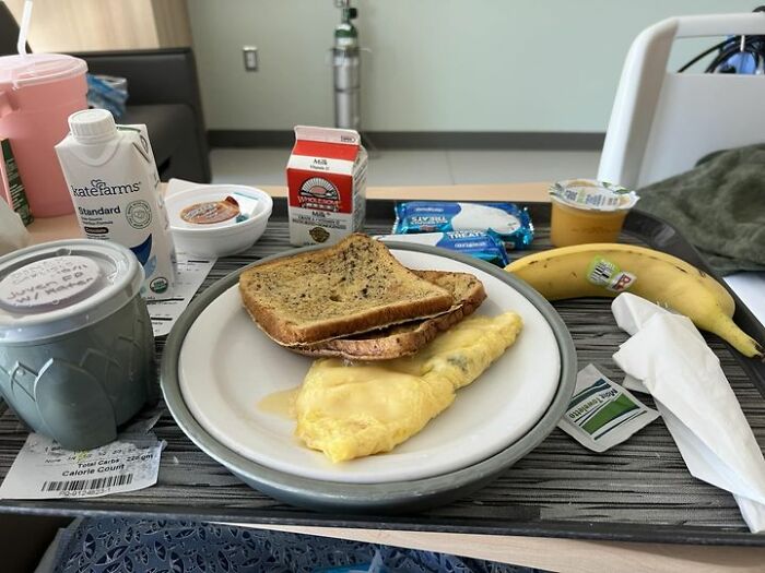 Hospital food tray featuring omelet, toast, banana, milk carton, and packaged snacks on a hospital bed table.
