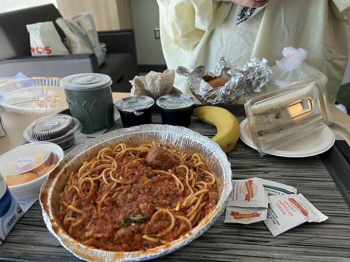 Hospital food example showing spaghetti with meat sauce, fruit cup, banana, sandwich, and drinks on a tray in a patient room.