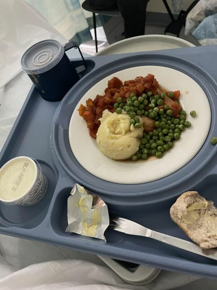 Hospital food tray with mashed potatoes, peas, vegetable stew, bread with butter, and a sealed drink cup in a healthcare setting.