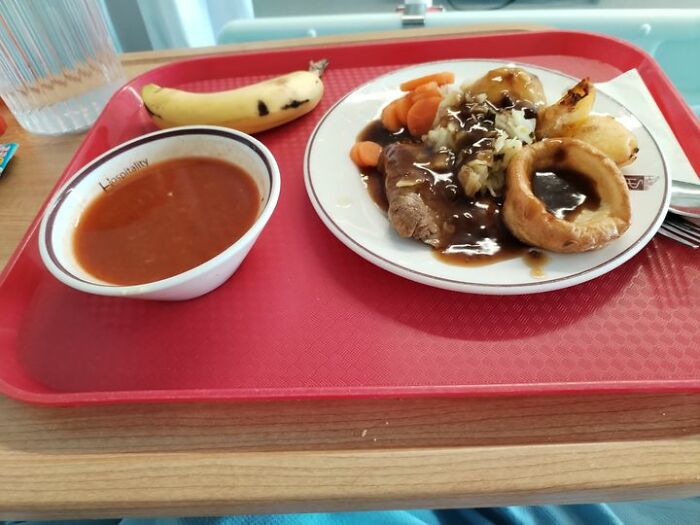 Hospital food tray with beef, gravy, vegetables, soup, banana, and water glass served on a red tray.