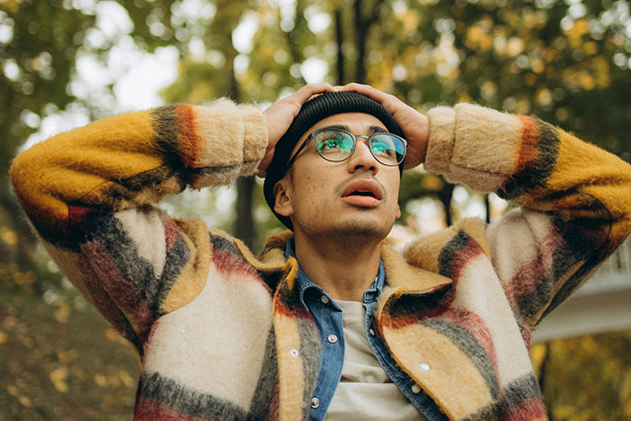Young man wearing glasses and a colorful jacket looks shocked outdoors, capturing moments that turned friends to strangers quickly.