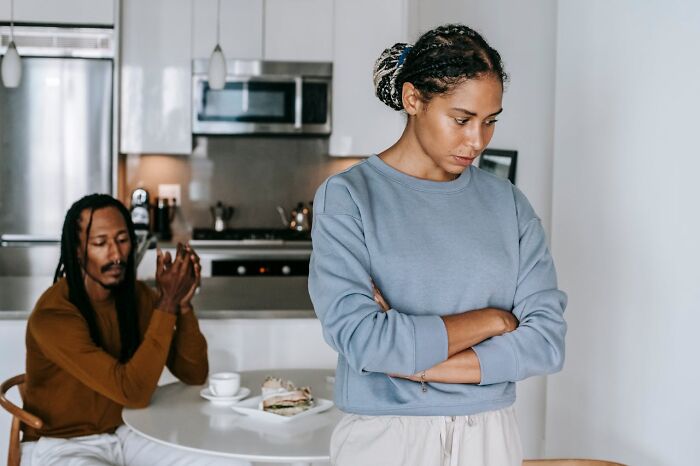 A woman looking upset with arms crossed while a man sits at a table behind her in a tense moment, toxic relationship.