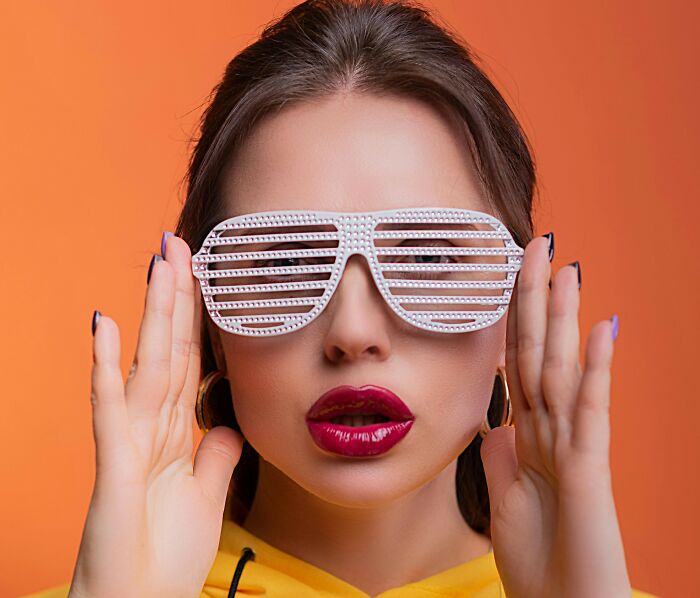 Young woman wearing white shutter shades and bold lipstick posing against an orange background, showcasing fleeting internet trends.