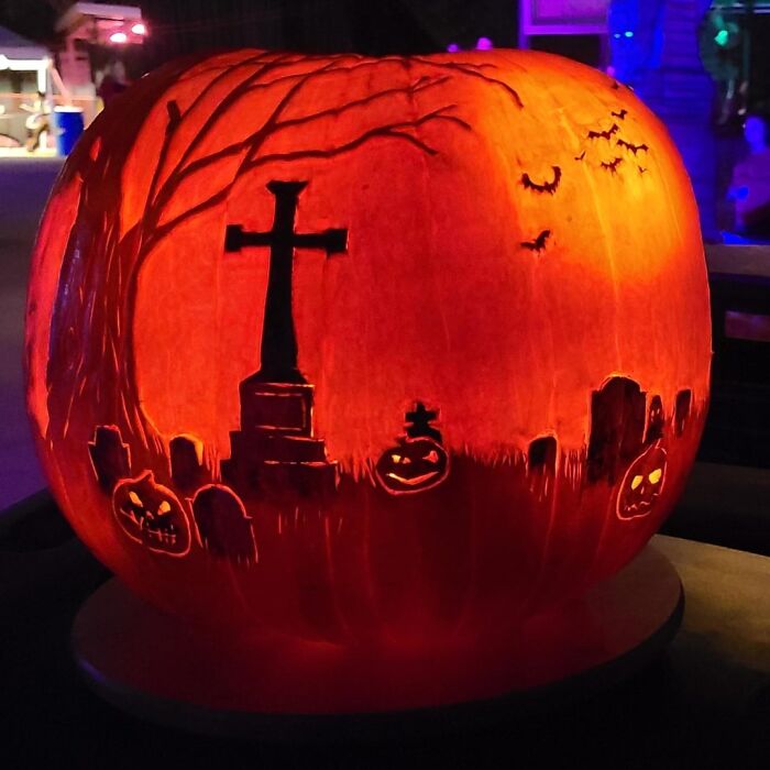 Glowing spooky pumpkin carving depicting a graveyard scene with tombstones, bats, jack-o-lanterns, and a large cross.