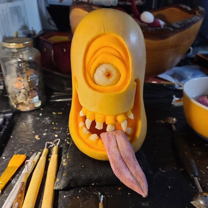 Carved quirky pumpkin with one eye, teeth, and a purple tongue on a messy crafting table with carving tools.