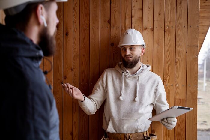 Construction worker wearing a hard hat and hoodie discussing mind-blowing coincidences while holding a tablet inside a wooden structure.