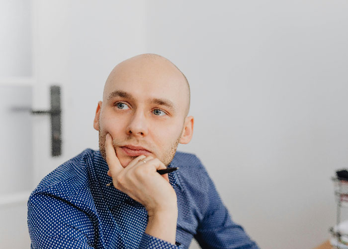 Young man in blue shirt thoughtfully holding pen, showing signs of being super smart and hiding intelligence well.