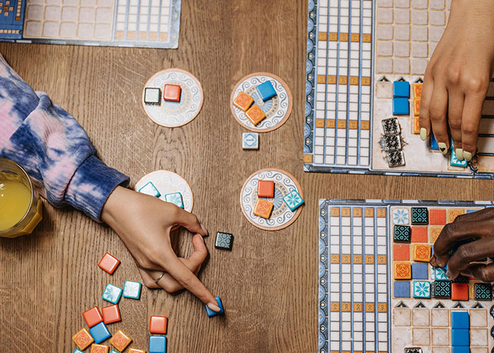 Two people playing a strategic board game with colorful tiles, showing indicators that the person is super smart and hides it well.