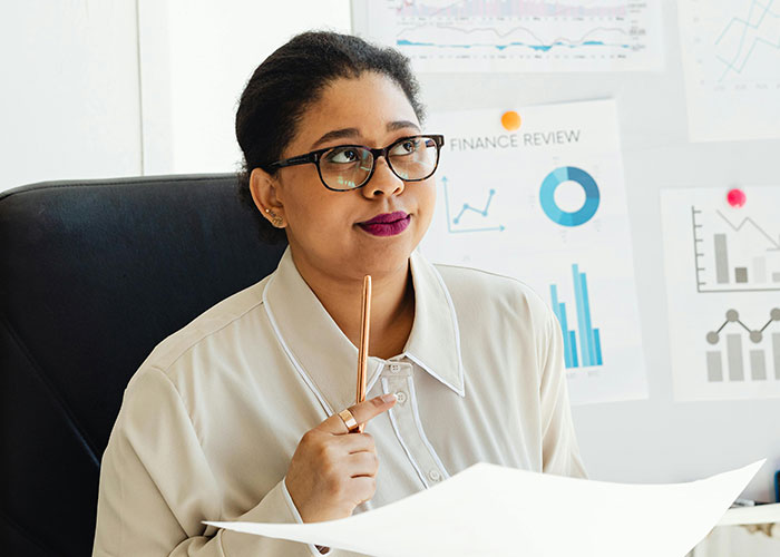 Thoughtful woman wearing glasses in office, reviewing charts and indicators showing signs of super smart intelligence.