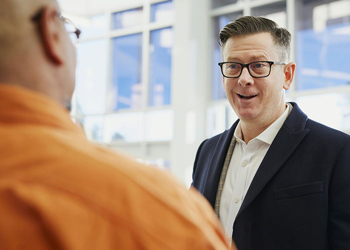 Man wearing glasses and a dark jacket talking to another person, illustrating indicators of a super smart person who hides it well.