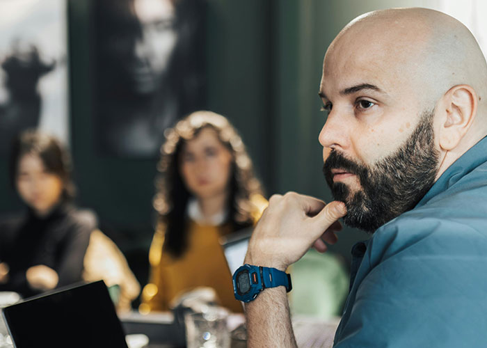 Bald man with a beard wearing a watch, attentively listening in a group discussion showing signs of hidden intelligence.