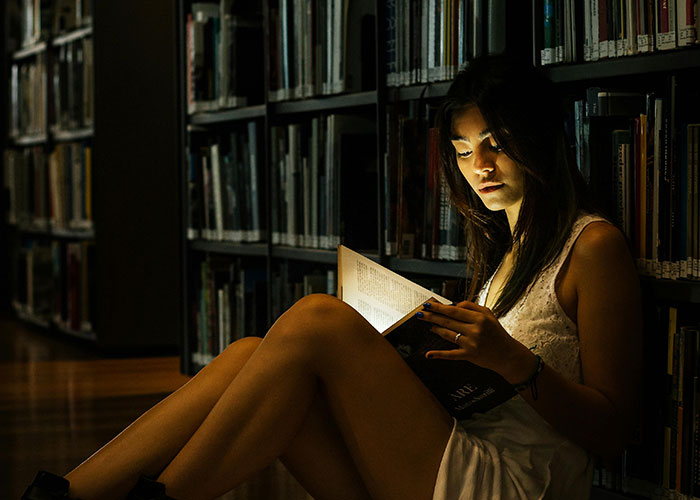 Young woman reading a book in a dimly lit library, illustrating indicators of a super smart person who hides it well.