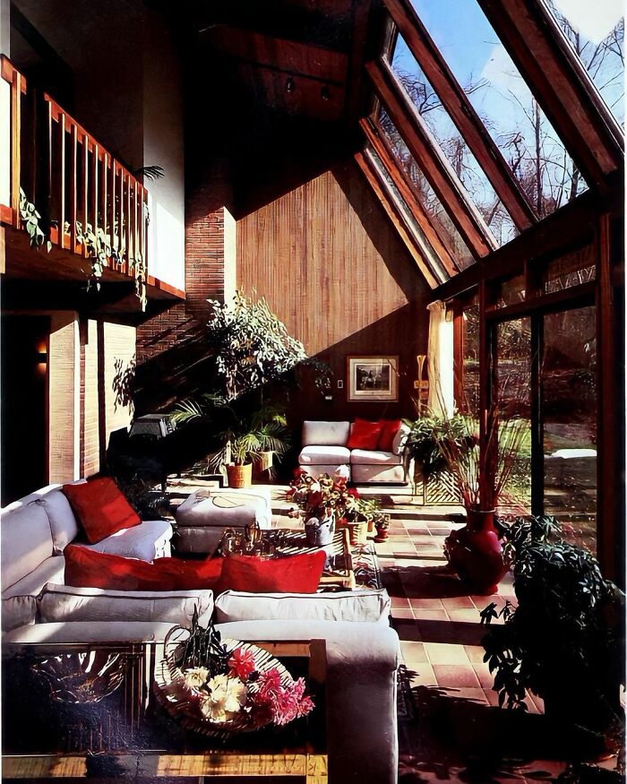 Mid-century interiors with large windows, wooden walls, and cozy seating filled with red cushions and green plants.