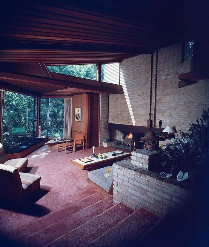 Mid-century interior with brick fireplace, wooden ceiling, large windows, and vintage furniture from the 50s to 80s decades.