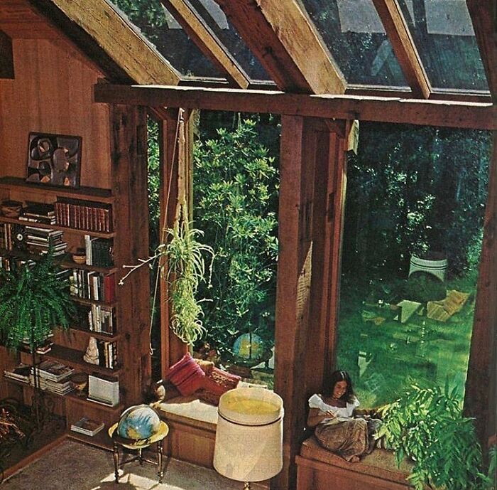 Cozy vintage interior from the ’70s featuring large windows, wooden beams, houseplants, and a woman reading by the window.