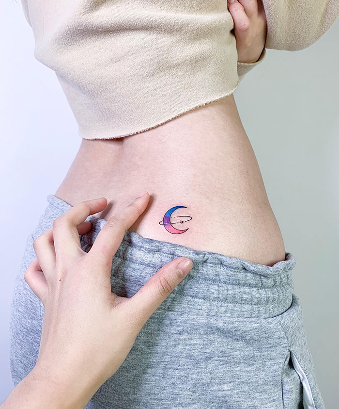 Small tattoo of a colorful crescent moon with planets on a person's side.