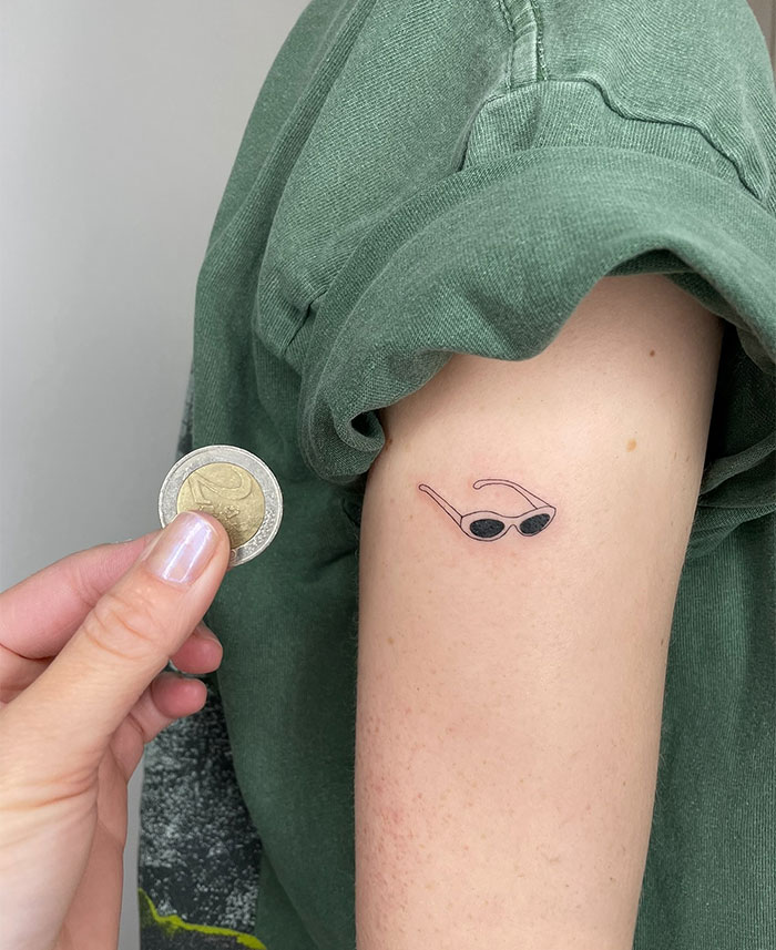 Small tattoo of sunglasses on an arm compared to a coin, illustrating tiny yet impactful tattoo ideas.